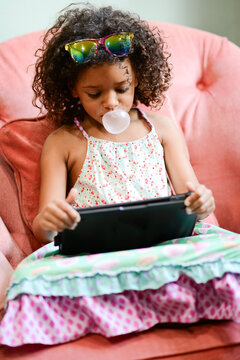 Young girl blowing a bubblegum bubble while using a tablet on a