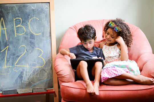 Two children sitting on a pink chair, using a tablet together, w - Powered by Adobe