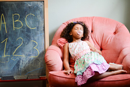 Young girl relaxing on a pink chair, blowing a bubble with bubbl