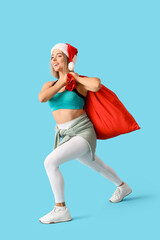 Obraz premium Sporty young woman in Christmas hat carrying Santa Claus bag on blue background