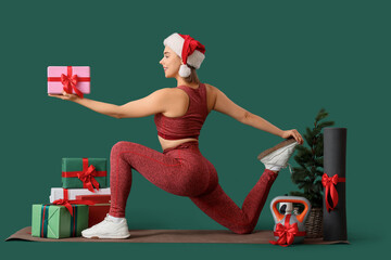 Sporty young woman with Christmas gift boxes, kettlebell and yoga mat exercising on green background