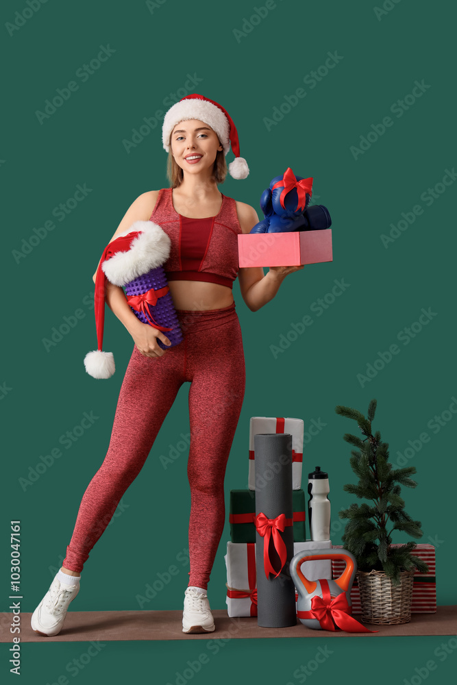 Poster Sporty young woman with Christmas presents and sports equipment as gifts on green background