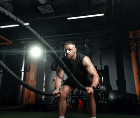 Fototapeta na wymiar Bodybuilder is engaged with battle ropes for muscle fitness in hall of gym. Training and endurance with intense workout for strength. Space for text