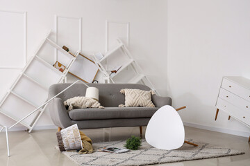 Interior of messy living room with grey sofa, overturned coffee table and shelving units after...