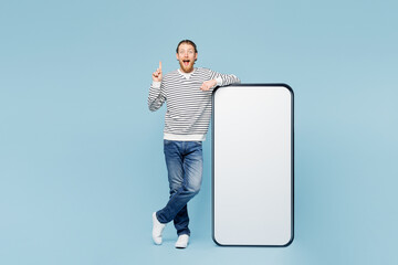 Full body young happy man wear striped sweatshirt casual clothes big huge blank screen mobile cell phone smartphone with area point index finger up isolated on plain blue background Lifestyle concept