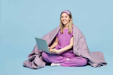 Full body calm young IT woman wear purple pyjamas jam sleep eye mask wrapped in duvet rest relax at home work hold use laptop pc computer isolated on plain blue background Good mood night nap concept