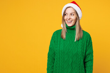 Merry shocked surprised young woman wear green warm cozy sweater Santa hat posing look aside on area mockup isolated on plain yellow background. Happy New Year celebration Christmas holiday concept.