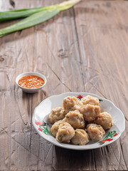 A plate of steamed Indonesian siomay served with a side of spicy dipping sauce. Fresh green onions are placed nearby, set on a rustic wooden table, creating a simple and appetizing presentation.
