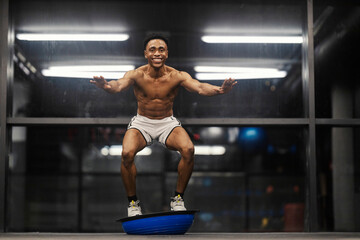 Full length of smiling strong powerful shirtless black sportsman squatting on bosu ball and...