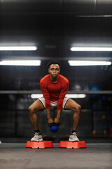 Young active strong muscular multicultural sportsman doing deep squats at gym with kettlebell and steppers.