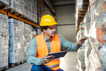 Interracial storage controller crouching next to goods and merchandise with tablet in hands.