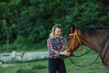 Obraz premium Happy blonde with horse in forest. Woman and a horse walking through the field during the day. Dressed in a plaid shirt and black leggings.