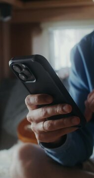 A young man with glasses smiles while using a mobile phone while sitting on a bed in a van. Close-up of his hands