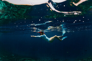 Young woman in blue wetsuit, fins and mask with snorkel swims underwater and looks at coral reef and fish near tropical island. Snorkeling and freediving activity during vacation.