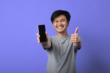 Fototapeta premium A young Asian man smiling, holding a smartphone in his left hand, while giving a thumbs-up with his right hand. He is wearing a gray t-shirt and standing against a purple background