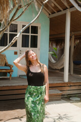 Portrait of a young blonde woman in swimsuit and light cape with tropical print standing against a background of bungalows with thatched roof during her vacation. Hotel on the island on ocean shore.