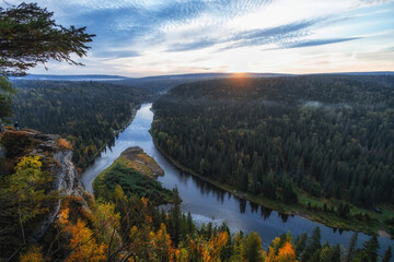 Obraz premium The sun sets behind rolling hills, casting a warm glow over a winding river embraced by vibrant autumn trees, creating a peaceful avenue of natureвЂ™s beauty