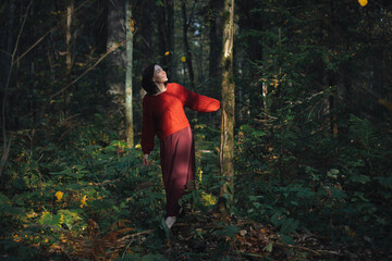 A woman in a red sweater gracefully stands in a vibrant forest, enjoying the sunlight filtering through the trees while observing the surrounding foliage in autumn