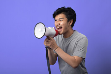 A young Asian man in a gray t-shirt stands against a purple background, holding a megaphone close to his mouth as if shouting, with an intense and energetic expression