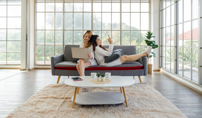 Two asian women sit on a couch, one using laptop computer and the other using mobile phone, in a bright room.