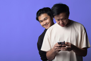 Fototapeta premium Two Asian men are standing against a purple background. The man in front is focused on his phone, while the man behind him is looking over his shoulder, seemingly curious about the phone