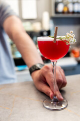 A hand holding a red cocktail garnished with a small white flower and a cinnamon stick, set against...