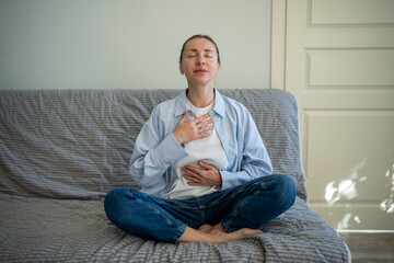 Calm woman sitting in lotus pose deep breathing during meditation at home. Self-help of middle aged...