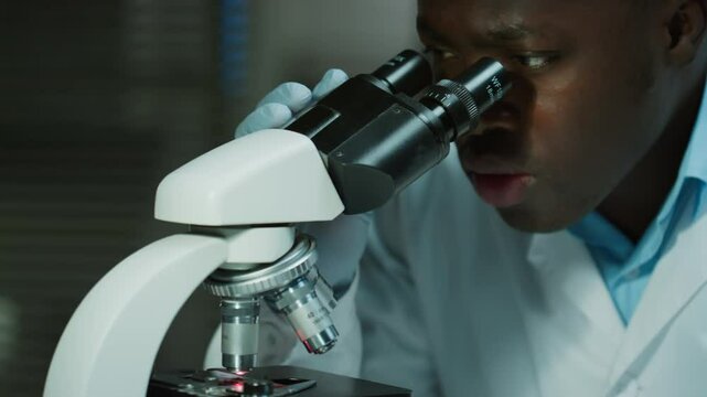 African American male scientist in medical gloves examining specimen under microscope while working in laboratory