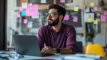 Obraz premium A man with a beard, wearing sunglasses and a purple shirt, sits at a desk in a modern office, looking away from his laptop