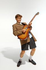 Young man dressed vintage attire and plays electric guitar passionately against white studio background. Concept of music, festivals, contemporary art, hobby, creativity. Ad