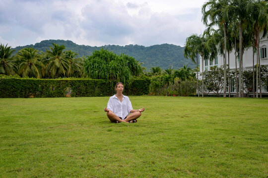Yoga of young woman meditating outdoors on the lawn under big tropical palms. Weekend Outdoor Classes takes place in the beautiful park. Beautiful tropical landscape