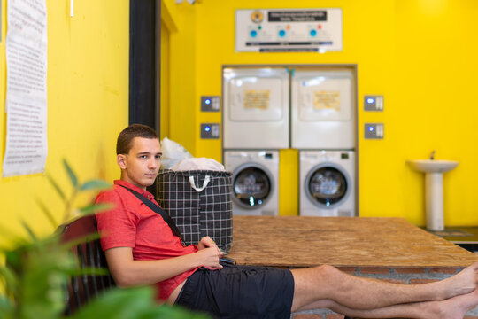 Bored young man sitting on the sofa by a washing machine and waiting for the laundry