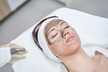Woman undergoing laser facial treatment with protective headband in modern skincare clinic.