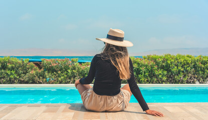 A woman wearing a straw hat sits on a pool deck