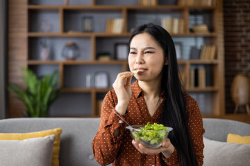 Asian woman eating salad with dissatisfied expression, contemplating diet. Seated on couch in contemporary interior setting, representing healthy lifestyle choices. Concept of wellness journey.