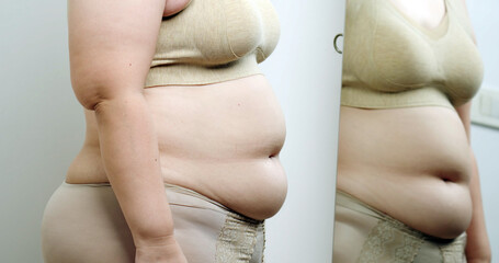Asian oversize woman in front of a mirror looks at her body. Excess belly fat, Unhealthy lifestyle, Diet concept.
