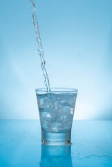 Water stream splashing on glass cup filled with water and ice on wet reflective blue surface
