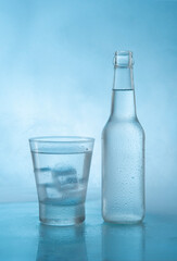 Frosted glass cup filled with water and ice next to glass bottle on wet reflective blue surface with steam