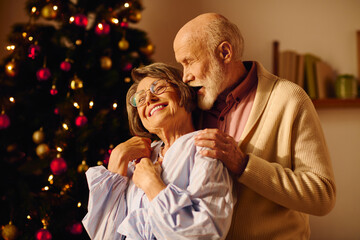 A senior couple joyfully embraces by their decorated Christmas tree at home.