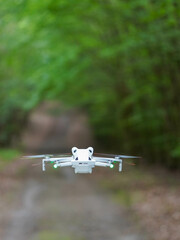 A small white drone is hovering in the air on a forest path, surrounded by green trees in the background