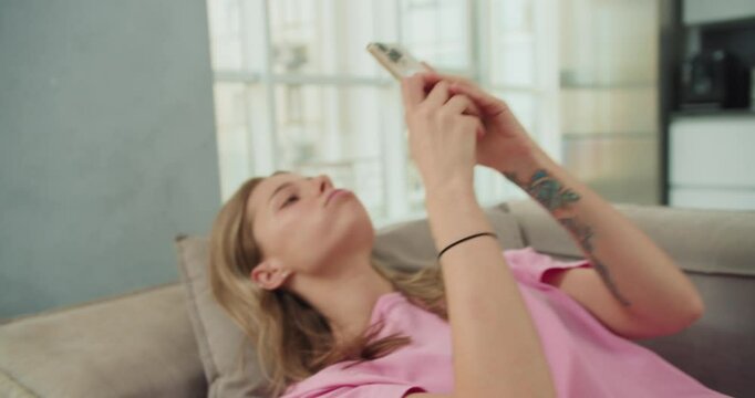 A young woman in a pink t-shirt laying on a sofa, holding her smartphone above her face, focusing intently as she scrolls through content, with a relaxed and casual atmosphere