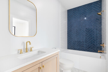 A white bathroom with a natural wood cabinet, gold faucet and mirror, and a shower with white...