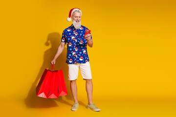 Fototapeta premium Full size photo of stylish modern santa smart phone shop bags empty space xmas winter print shirt isolated on yellow color background