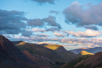 Mountain and hill tops illuminated by sunset light. Rocks silhouettes and gold sunrise colors. Vivid golden sunlight on hills under clouds of sunset tones in blue sky. Shadows of clouds on rocky ridge