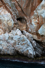 Brown colored Basalt sheer rocks  cuted by cracks due geology and by constant wind and temperature fluctuations. This cliff  is riddled with deep caves. Cliff downs to  blue water of the sea.