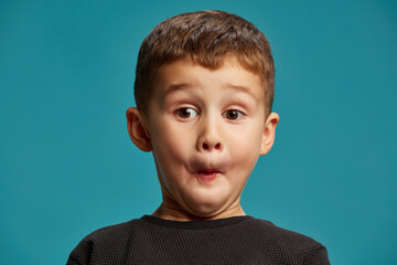 Boy makes comical expression with puffed cheeks, his eyes wide open in surprise against blue studio background. Childhood curiosity. Concept of facial-expressions, positive and negative emotions. Ad