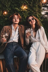 Fototapeta premium A man and a woman are sitting on a bench, holding wine glasses and smiling. The man is wearing a brown jacket and the woman is wearing a white shirt and white pants. Scene is happy and relaxed