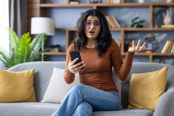 Obraz premium Portrait of a disappointed young Muslim woman at home, sitting on the couch at home, holding a phone in her hand, spreading her hand and looking upset at the camera