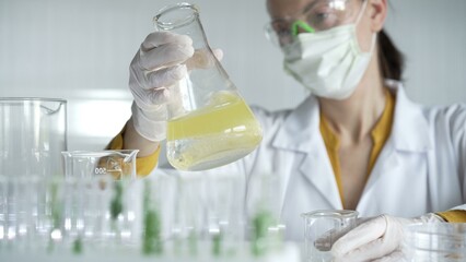 Woman scientist wearing white protective gloves is shaking up a yellow chemical solution inside an...