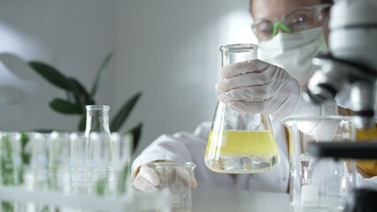 Woman scientist wearing a lab coat, white gloves, and a mask is pouring a yellow oily liquid from one beaker to another in a laboratory setting, close up. Science and medicine